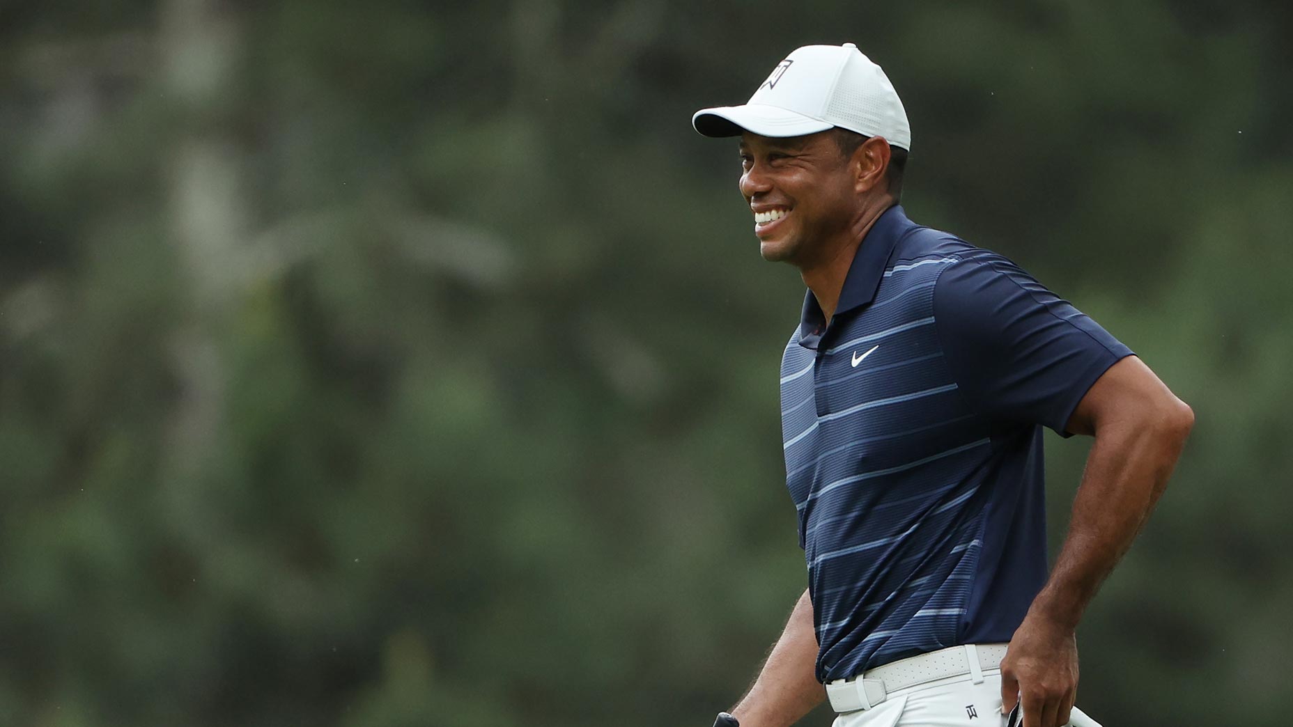 tiger woods smiles in a white hat and blue shirt from a fairway at the masters tournament